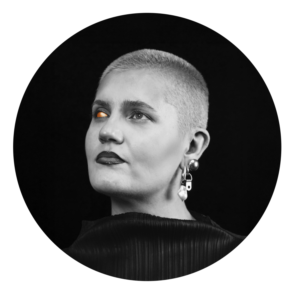 Black and white portrait of Aksana Berdnikova. Aksana is an Eastern European woman with shaved, bleached hair and chunky earrings. Her left eye is prosthetic and gold.